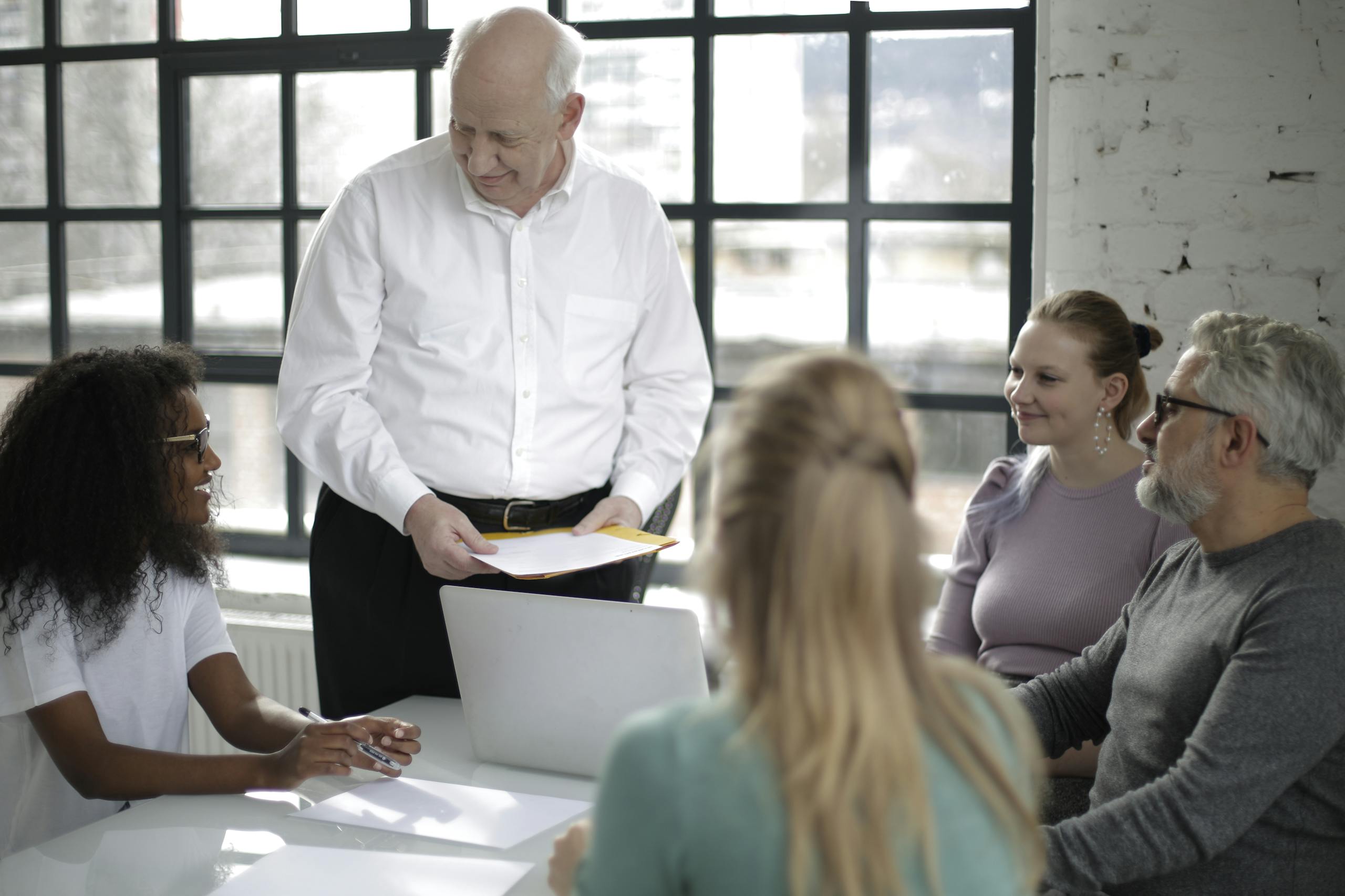 Positive multiracial colleagues of different ages gathering at table in modern workspace while older man presentation project